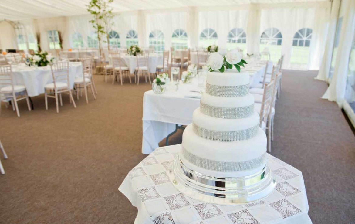 Wedding marquee with tables set up for dining and the 4 tier white wedding cake on the corner table beside the top table